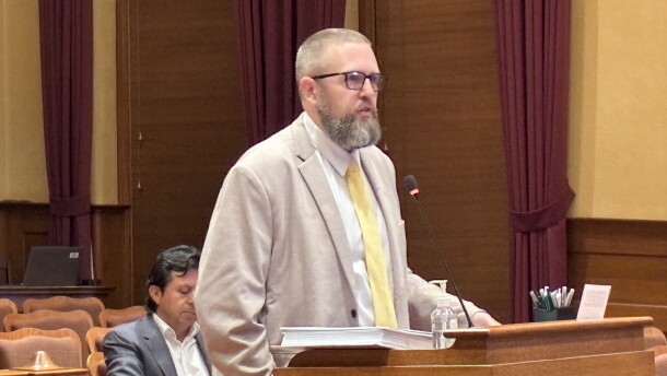 A bearded man in a blazer and tie speaks at a podium in a courtroom, with another man sitting behind him.