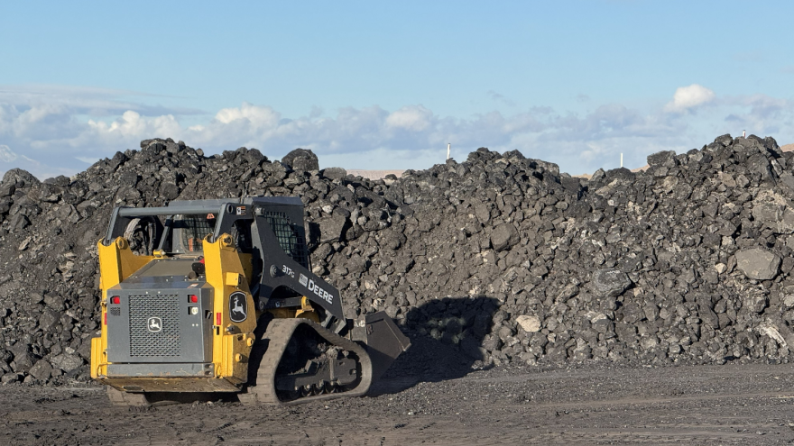 A construction vehicle with a scooping implement is in front of a pile of coal taller than the vehicle that stretches past the edges of the photo.