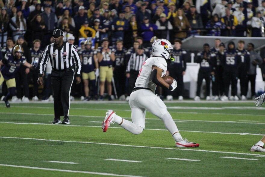 A football player in a white uniform runs with the ball during a game, with a referee and opposing players in dark uniforms visible in the background, and a crowd watching from the stands.