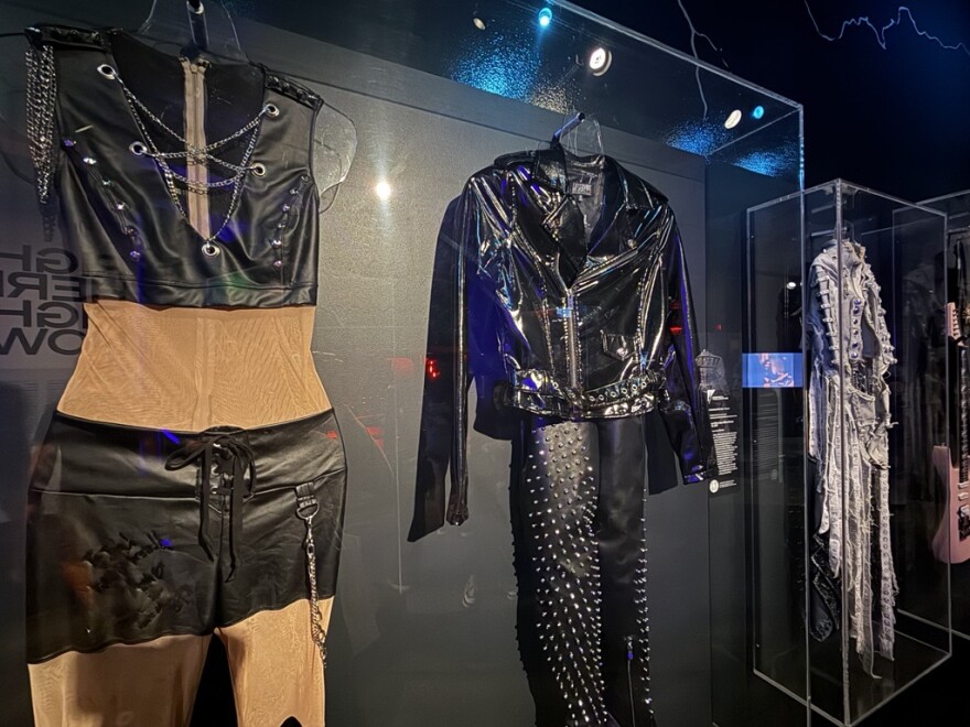 Two stage outfits hang side by side in a glass museum case: a black leather vest and shorts with silver chain lacing, and a black leather jacket with metal-studded pants. On the right, a silver and denim stage outfit hangs in another glass museum case.
