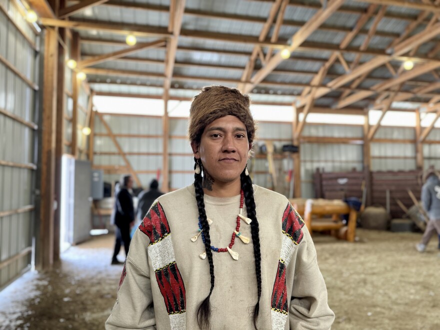 A man with braids and a fur hat poses for a picture inside a barn.
