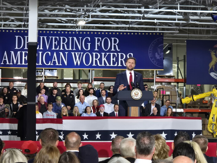 JD Vance speaks at a lectern in front of a blue banner that reads "DELIVERING FOR AMERICAN WORKERS."