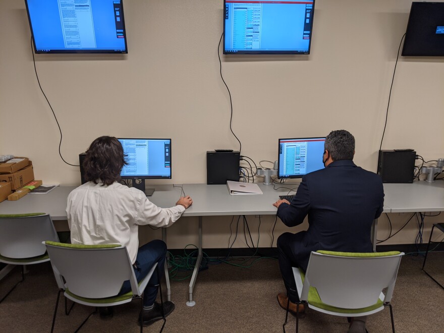 Two men sit in front of computers that are attached to monitors overhead.