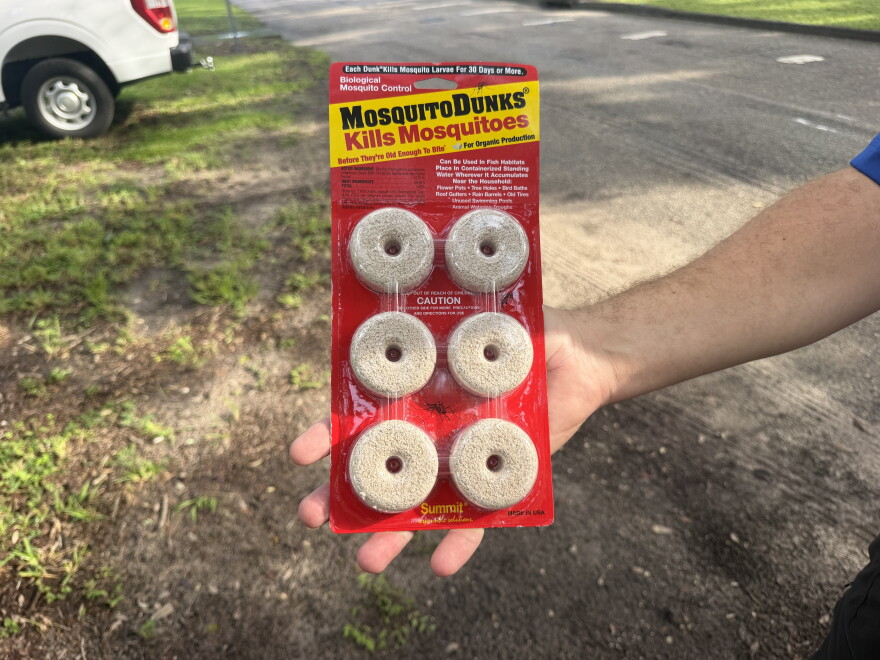A hand is holding a red and yellow package that reads, "MosquitoDunks, Kills Mosquitoes." Inside the clear packaging there are six beige, donut-like pellets. The is grass and a dirt road in the background.