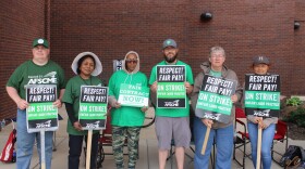 A group of 6 adults stand together in green shirts and holding green signs on a concrete pad. They are protesting.