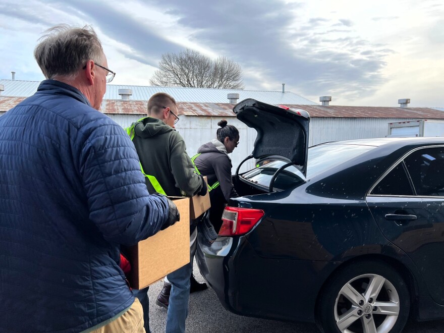 A line of volunteers carry cardboard boxes to a car with its trunk open.