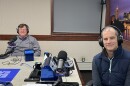 Two men wearing headphones sit at a table in a radio talk studio: a man at left has short brown hair and is wearing glasses and a grey button-down shirt; a man at right has short dark hair and is wearing a navy puffy vest over a navy long-sleeved shirt.