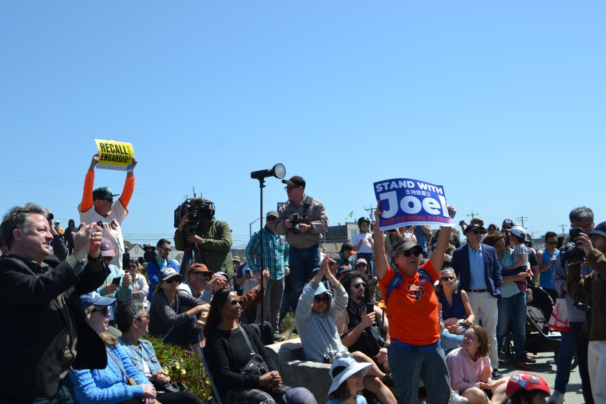A man in a Giants jersey holds a "Recall Engardio" sign up behind a woman holding a "Stand With Joel" sign
