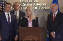 Blonde woman standing at a podium and microphone with a man in dark suit to the left, and an older bald man in a dark suit to his right