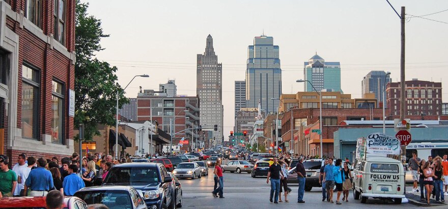 While the sky is still light, a large street in the middle of Kansas City is crowded with cars and people.
