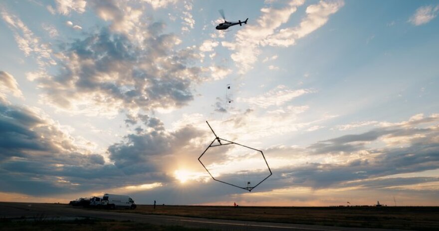 A thin hexagon dangles from a helicopter over an empty field. The sun peeks through the clouds, with vans parked in the left of the image.