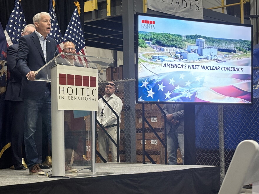 A man stands at a clear podium labeled "HOLTEC INTERNATIONAL" while speaking into a microphone on a stage. To his right, a large digital monitor displays an aerial view of a nuclear power plant with the text "AMERICA'S FIRST NUCLEAR COMEBACK" overlaid on an American flag design. Additional individuals stand in the background behind a chain-link fence, and an American flag is partially visible to the left.
