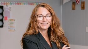 Red headed woman wearing glasses, standing in a classroom with her arms folded, wearing a blue blazer
