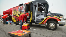 A man wearing a red jacket and red ball cap sets down a yellow and red ice chest from a large, colorful van that has Kansas City Chiefs wording and logos printed on it.