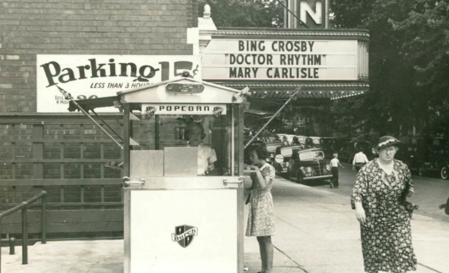 Image of the Irvin Theater in Bloomington. A woman buys popcorn from the stand outside. Another older woman in a long dress and hat walks by. The marquee sign is for Bing Crosby "Dr Rhythm" and Mary Carlisle.