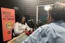 African American woman sits with headphones on near microphone in front of WUSF Your Spot for Local sign. A man's back with headphones over his ears is also seen sitting across from her next to another microphone.