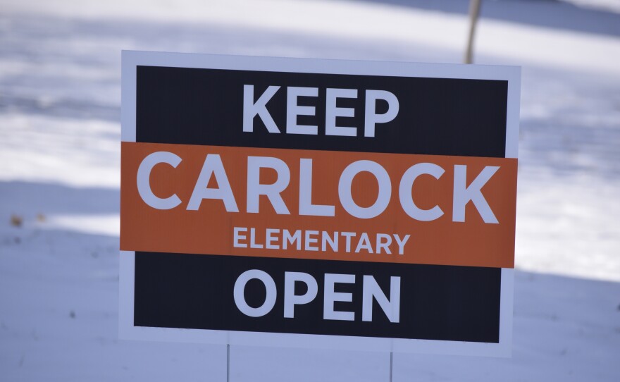 A black yard sign says, "Keep Carlock Elementary Open." There is snow in the background.