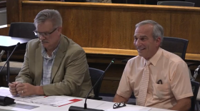 Two men sitting at a table behind tall mics. The man to the left is wearing a green jacket and glasses, and the man to the right is wearing a light orange dress shirt and light orange tie. 