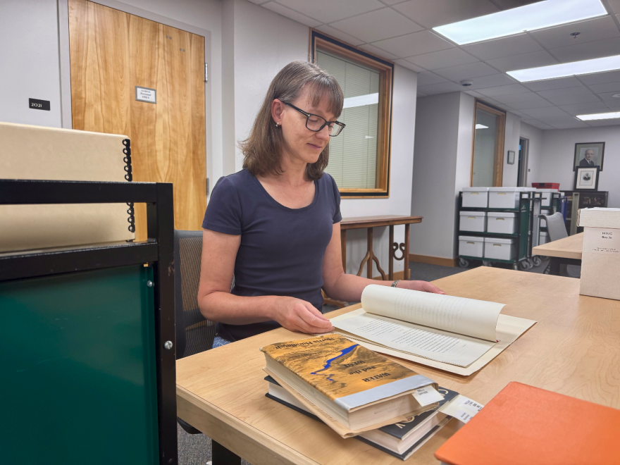 A woman sits at a table flipping through old papers with a few books off to the side. There are several carts full of boxes in the room nearby.