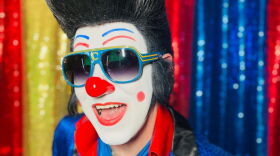 A man with clown face make up also looks like Elvis Presely with a fake black updo wig and a red nose. He is smiling.