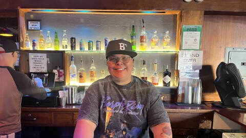 Patty stands behind the bar at the White Horse wearing an Oakland Ballers cap and a Prince t-shirt, smiling.