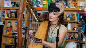 A woman in vintage 1940s clothing sings and plays a harp while seated behind an office desk with shelves of music paraphernalia behind her.