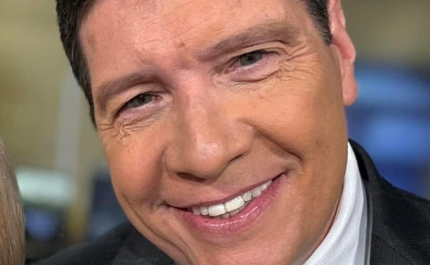 A smiling man with short dark hair wears a dark grey blazer, white button-down shirt and red and blue striped tie.