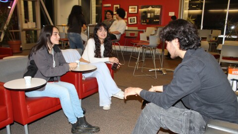 In the foreground, two people with long dark hair sit on red chairs across from another person with shorter dark hair. In the background, three other college students chat around a circular table.