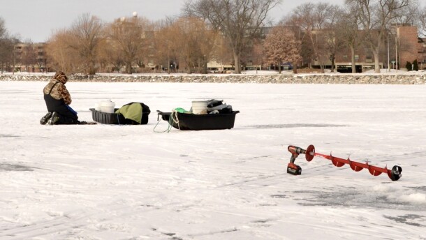 A man kneels on a frozen body of water to measure ice thickness. A red ice auger sits on the ice nearby.