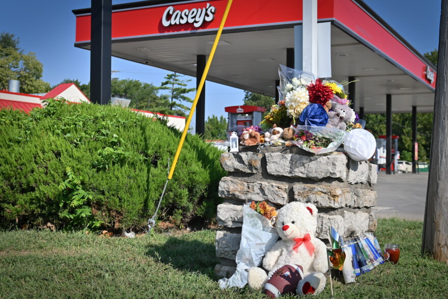 Outdoor photo shows a collection of flowers, candles, a football and other tributes lying around a concrete block. In the background is a gas station with a sign that reads "Casey's."