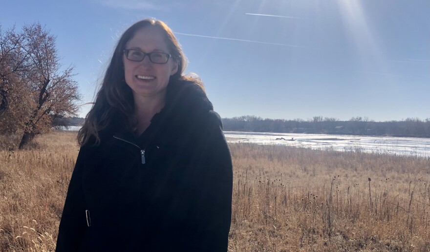 A woman in a black coat and glasses stands in front of a river, with tall brown grass behind her.