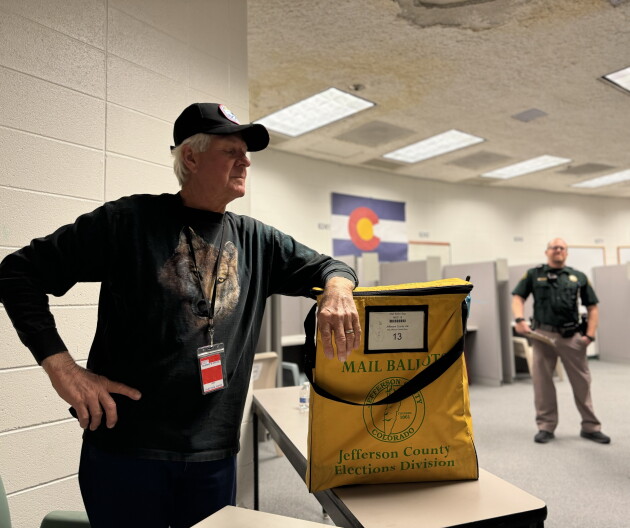 An election volunteer in the Jefferson County Jail stands behind a folding table with a yellow ballot bag. 