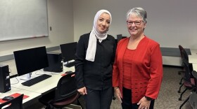 Two women standing in a classroom between rows of students desks and computers. The women on the left is wearing a dark suit with white hijab, the woman on the left is wearing a red top and jacket with blank pants.