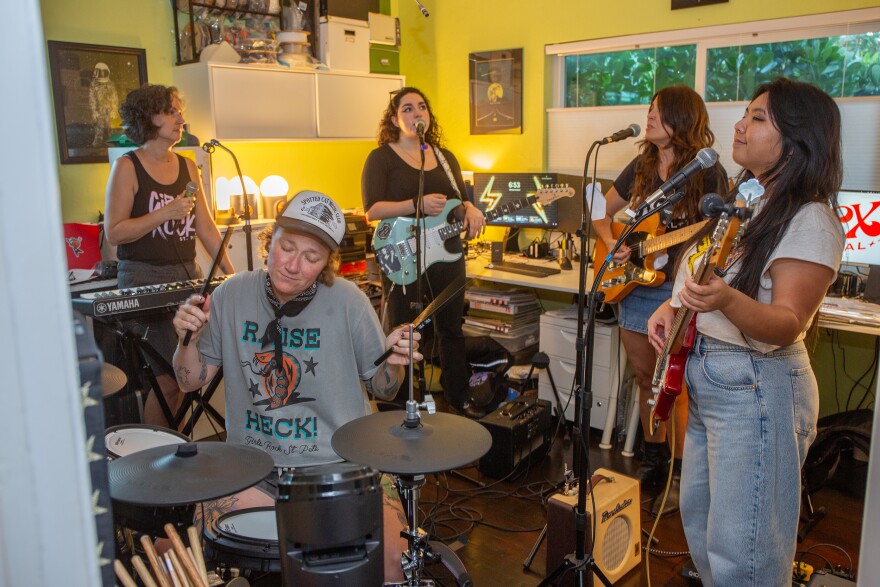 Five women in casual clothes cram inside a small bedroom with green walls while they practice as a band.