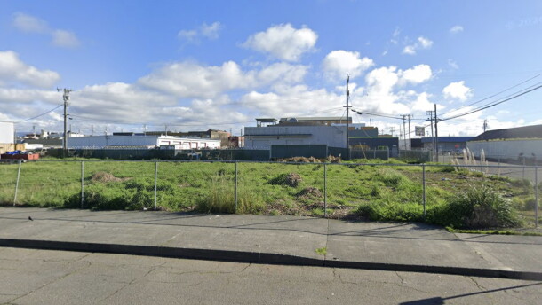 An empty site is covered with green grass and lined by chainlink fence. It's surrounded by white industrial buildings. 