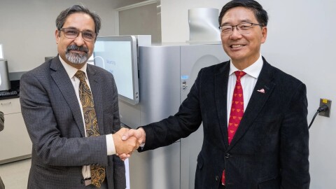 Two men dressed in suits and ties shake hands as they stand in front of a large silver machine, about as big as a refrigerator. 