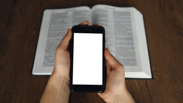 A woman's hands hold and a smart phone touch with a blurred Bible behind her hands
