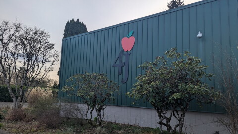 Green outdoor wall with 4J and an apple logo on it. in the foreground bushes and a sidewalk.