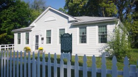 A white wooden house sits behind a tall white fence. Trees surround the building, with a single bush straddling its side. A sign sits just in front of the building, informing visitors of the abolitionist history of the site. 
