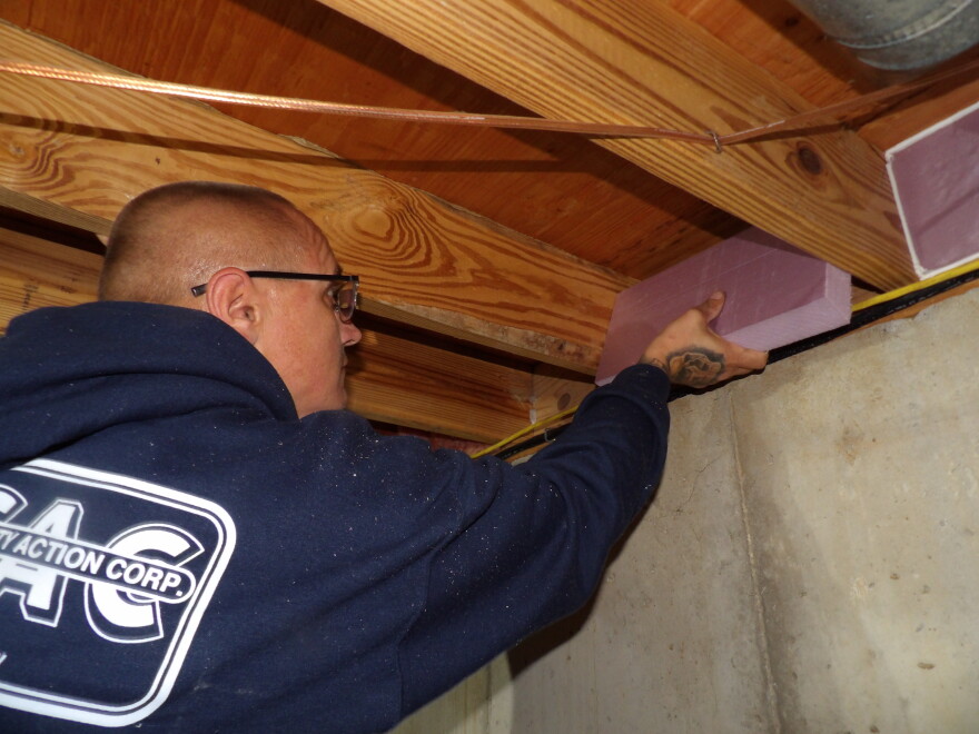 An over-the-shoulder image of a bald man in a navy blue hoodie placing a piece of purple foam insulation in between the beams of a ceiling. 