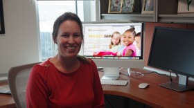 A woman in a red blouse sits in a desk chair, smiling towards the camera. A computer monitor behind her displays an advertisement for Project SHINE, with a young girl in a pink dress seen smiling in the ad. 