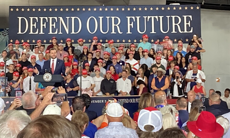 Man speaks at podium in front of a crowd and a sign saying Defend our Future