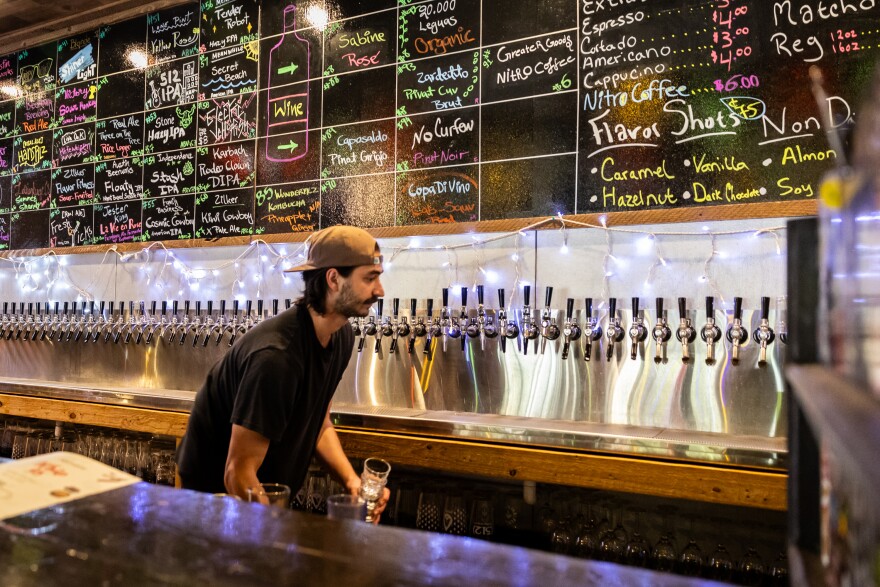 A bartender in a black shirt with a backwards baseball cap is holding a glass as he approaches a long line of draft beer taps. A stainless steel backsplash is behind the beer taps. Above, a chalkboard shows upcoming events and a coffee menu. 