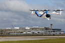 A white and blue electric aircraft flies at Tampa International Airport during a demonstration flight, with six propellers visible.