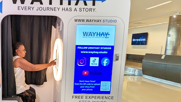 A woman sits inside a video booth and reaches out with one arm to tap the screen in front of her. The booth is decorated with blue and black arrows and text against a white background. "WAYHAY" and the tagline "Every Journey Has A Story" are at the top of the booth, over the doorway to the compartment where the woman is sitting.