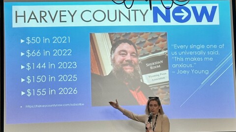 A woman stands in front of a PowerPoint screen with her hand extended, speaking into a microphone. An image of a bearded man and some info about “Harvey County Now” is displayed behind her.
