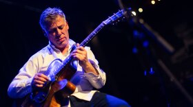 A man plays a hollow body guitar on a stage against blue light and a dark background.