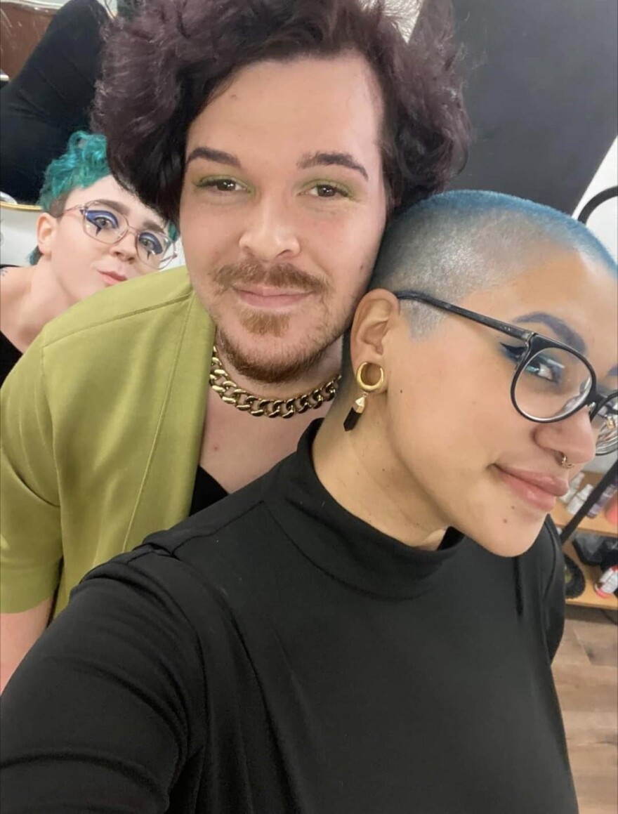 Ash Ratts a white person with short blue wavy hair stands behind Sydney Eckhoff and Rizza Benton. Sydney, a white-non binary person with a goatee and short wavy light brown hair is smiling and wearing a mustard cardigan with a black undershirt. Sydney is standing next to Benton. Benton is wearing a long sleeve black turtle neck and round glasses. Their hair is shaved with blue tips on the end. Benton is a light skin Black non-binary person. 