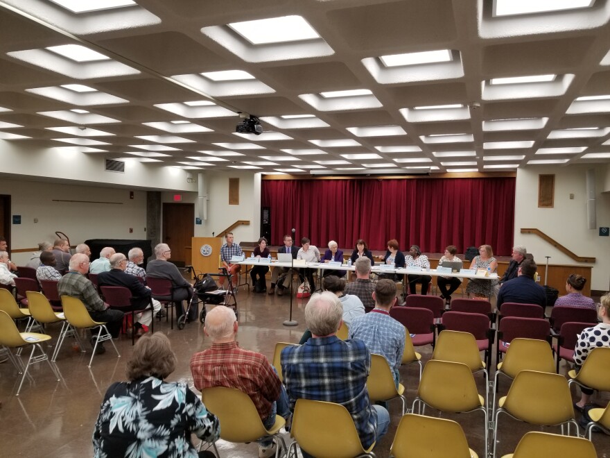 Rows of seated seniors face a panel of also seated people behind a table in a basement auditorium