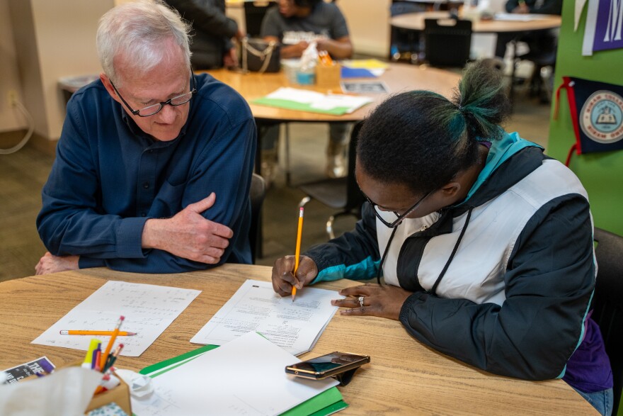 Man sits a table with woman helping her work on a paper.